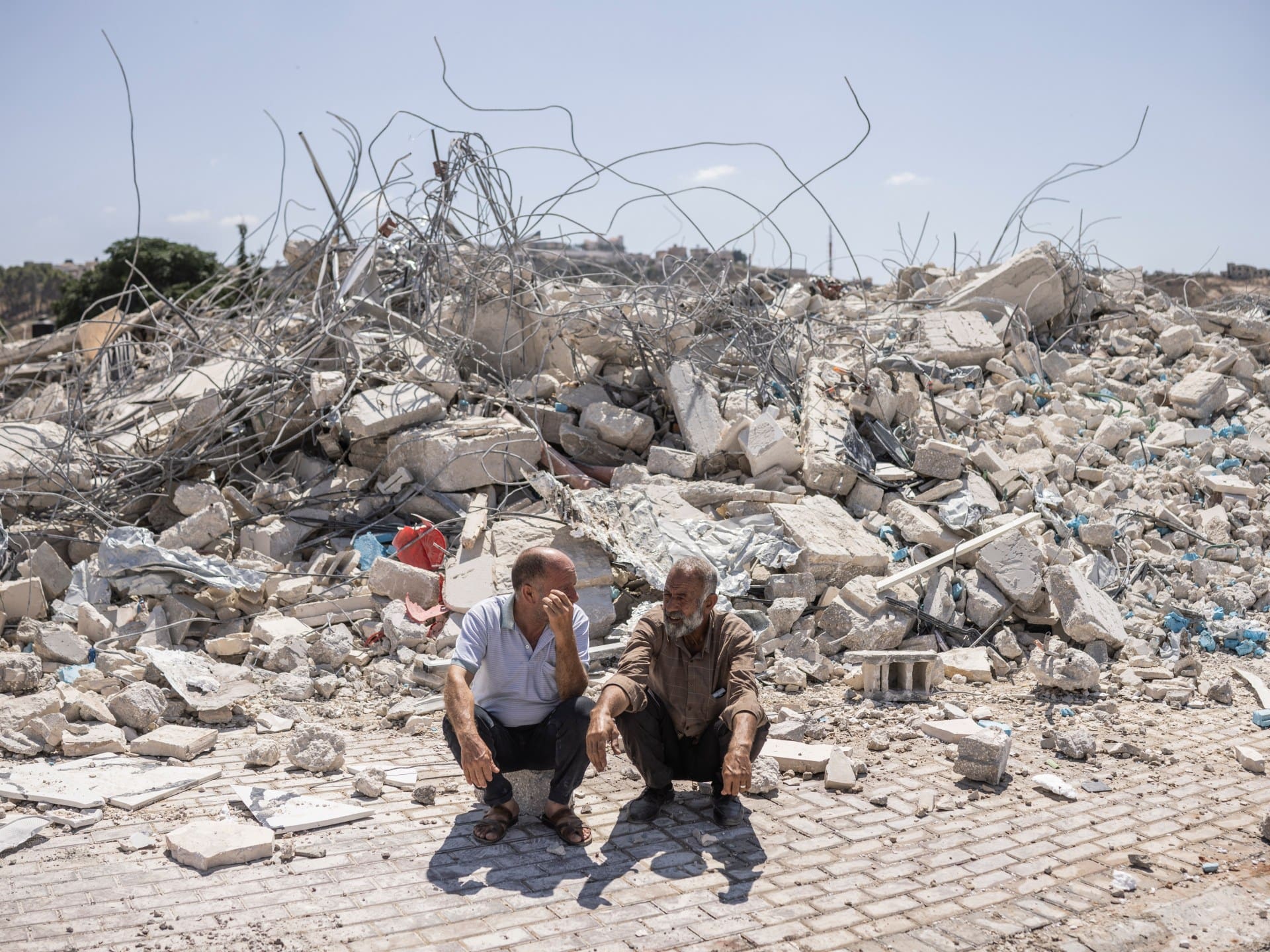 Two Palestinian men crouch in front of the rubble remains of a building containing shops and apartments that were demolished by Israeli forces, citing the lack of a building permit, in Arraba, south of the Palestinian city of Jenin in the northern Israeli-occupied West Bank, on July 27, 2026.