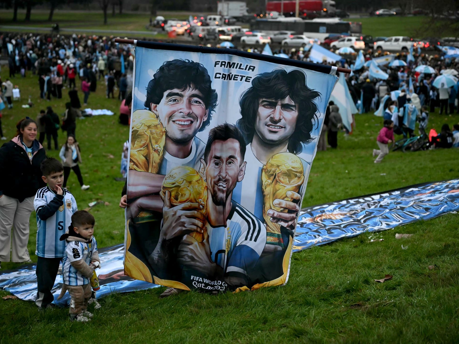 Fans stand beside a banner depicting Argentine football legends Diego Maradona, Mario Kempes and Lionel Messi holding FIFA World Cup trophies as supporters gather near the Argentine Football Association (AFA) training camp in Ezeiza, Buenos Aires Province, on July 20, 2026, while awaiting the arrival of Argentina's national football team from the United States following its defeat in the 2026 FIFA World Cup final. (Photo by Luis ROBAYO / AFP)